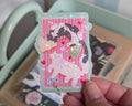 A hand holding a die-cut sticker featuring two cute illustrated cats on a pink striped background with small hearts, stars, books and a games controller. The sticker has a holographic glitter overlay.
