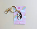 A glitter keychain featuring a black and white cat with gold glitter spots, resting on its pink backing card with rainbows and clouds.