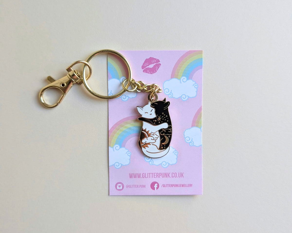 A glitter keychain featuring a black and white cat with gold glitter spots, resting on its pink backing card with rainbows and clouds.