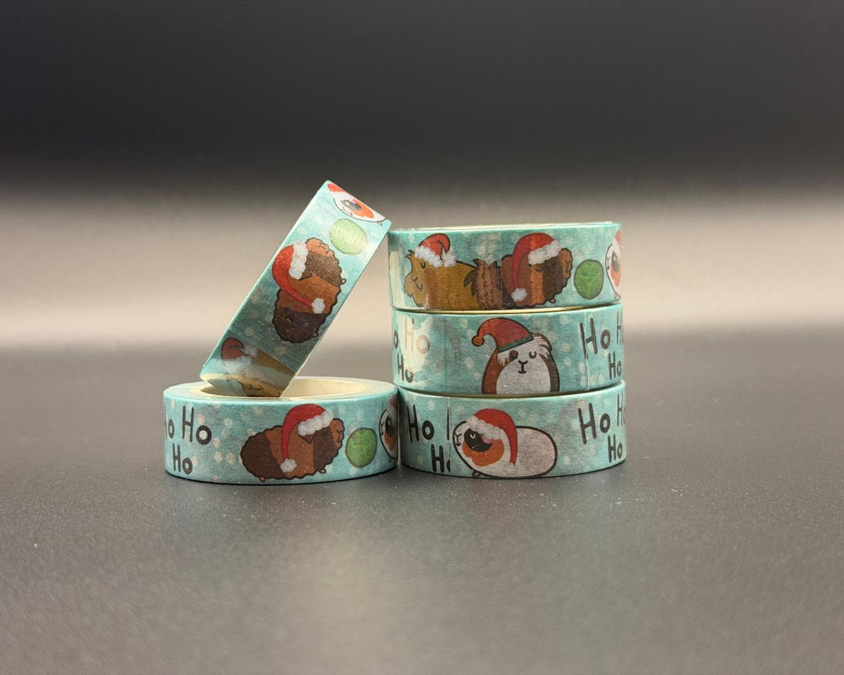 Decorative washi tapes featuring a Christmas guinea pig pattern and the words, 'ho ho ho'. The background of the tape is a light blue with white dots representing snow. There are also Brussel sprouts. 