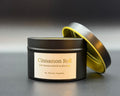 A black metal candle tin with a cream label reading 'Cinnamon Roll' and 'By Thistle Candles,' shown with the lid propped behind it on a dark gray background.