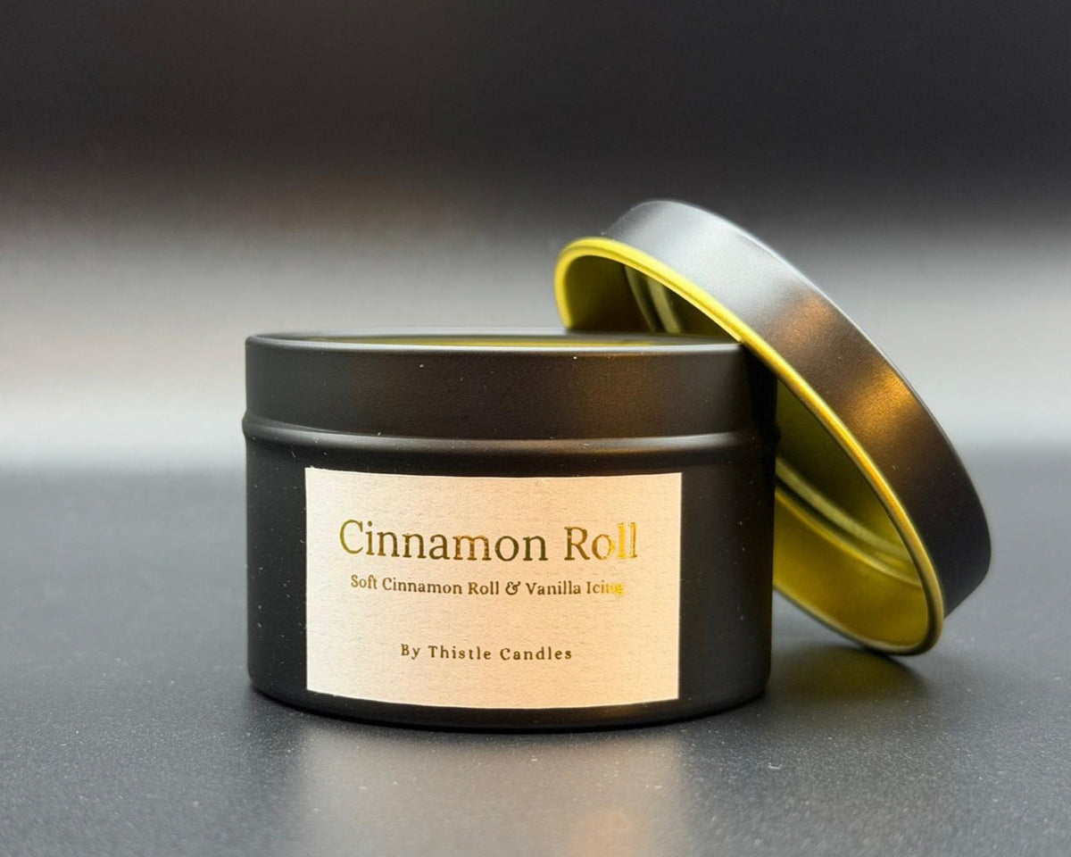 A black metal candle tin with a cream label reading 'Cinnamon Roll' and 'By Thistle Candles,' shown with the lid propped behind it on a dark gray background.