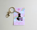 A black cat enamel pin sitting on a stack of purple books, attached to a gold-tone keyring and displayed on a pink backing card with a rainbow and clouds.