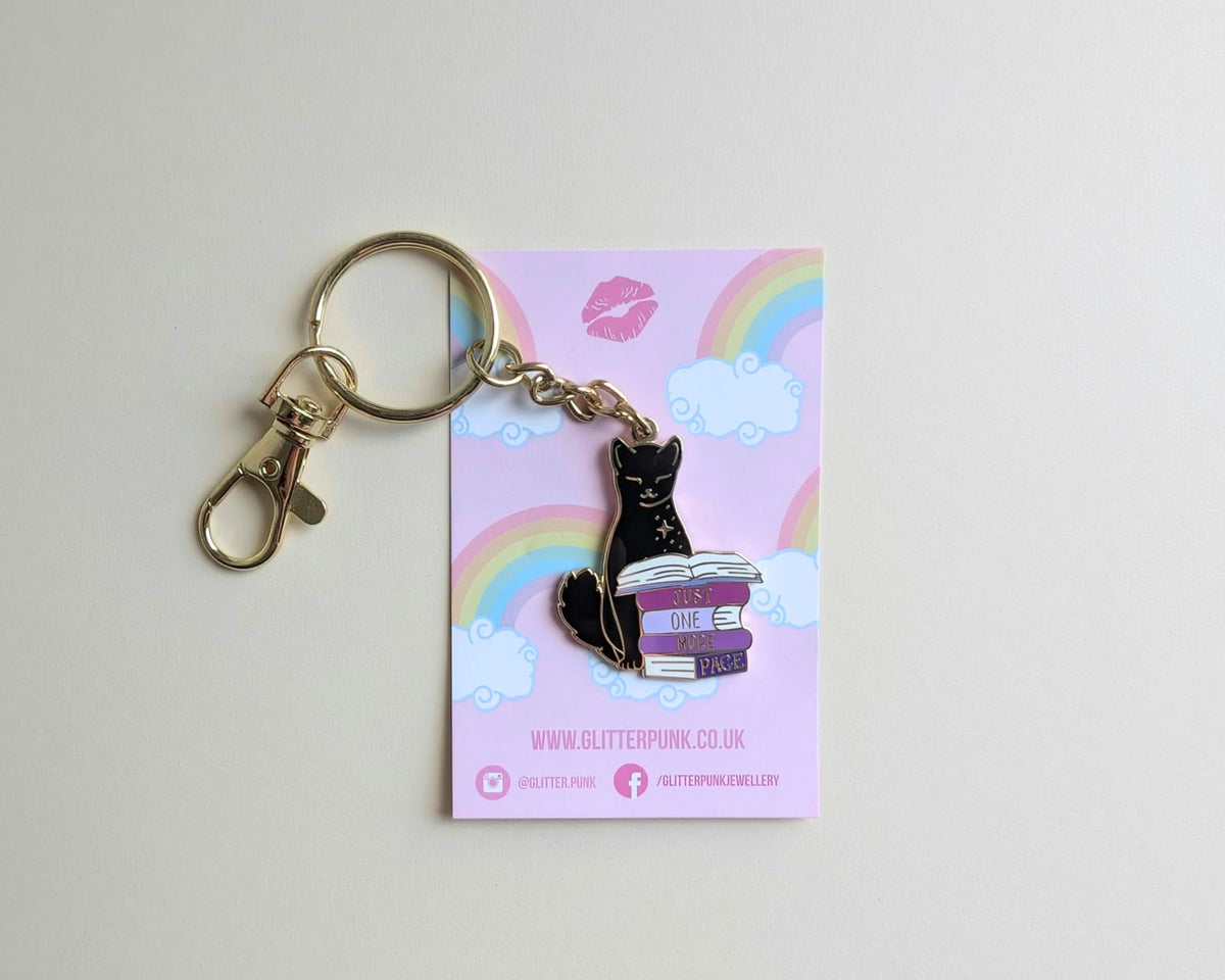 A black cat enamel pin sitting on a stack of purple books, attached to a gold-tone keyring and displayed on a pink backing card with a rainbow and clouds.