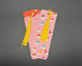 Pink paper bookmark with red, green, orange and pink Christmas hats. Has rounded corners and a yellow fabric tassel. 