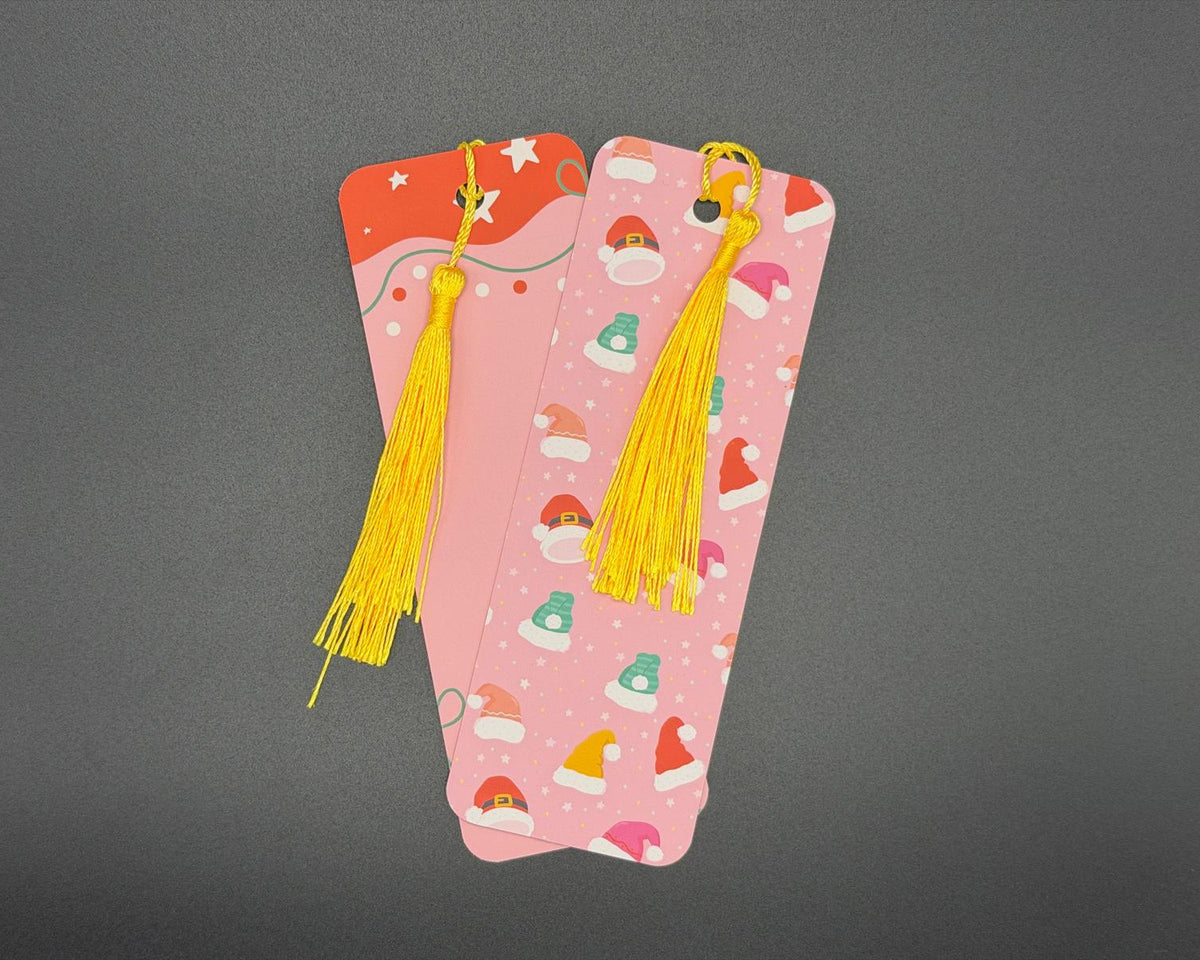 Pink paper bookmark with red, green, orange and pink Christmas hats. Has rounded corners and a yellow fabric tassel. 