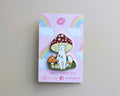 A cute enamel pin of a white cat sitting under a red-and-white spotted mushroom, displayed on its pink backing card with rainbow graphics. The backing card is branded with 'GLITTERPUNK'.