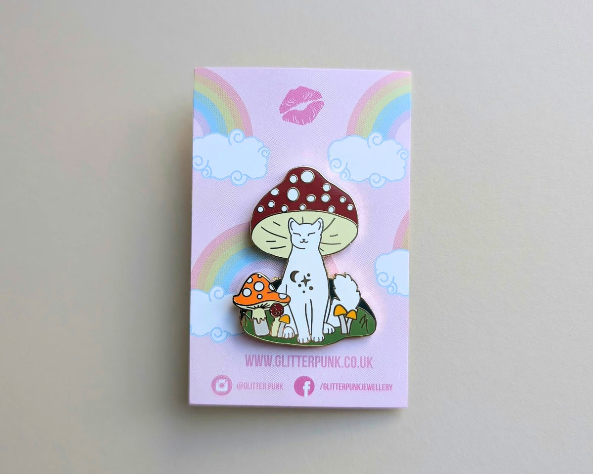 A cute enamel pin of a white cat sitting under a red-and-white spotted mushroom, displayed on its pink backing card with rainbow graphics. The backing card is branded with 'GLITTERPUNK'.