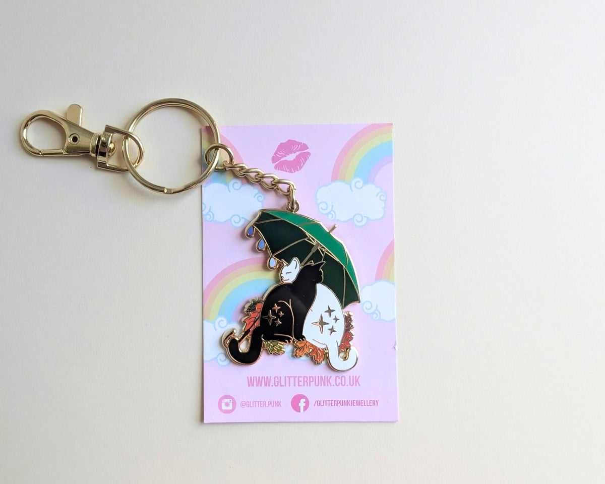 A Glitter Punk enamel pin of a black and white cat with a green umbrella, attached to a gold keyring and displayed on its pink backing card.