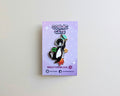 A cat enamel pin on a lavender backing card, photographed against a plain cream wall. The pin depicts a black-and-white cat wearing a green space helmet and a grey rocket, with orange flames.