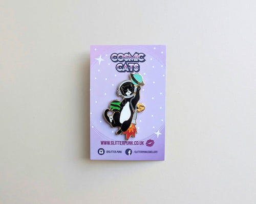 A cat enamel pin on a lavender backing card, photographed against a plain cream wall. The pin depicts a black-and-white cat wearing a green space helmet and a grey rocket, with orange flames.
