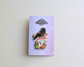A raven enamel pin displayed on its lavender backing card, which features the brand name 'Corvid Collection'. The pin depicts a black raven perched on a red-and-white mushroom next to a gold star and autumn leaves.