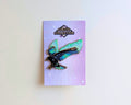 A raven enamel pin with teal, green, and black wings and star details, mounted on a lavender backing card from Glitter Punk's Corvid Collection, displayed against a plain off-white wall.