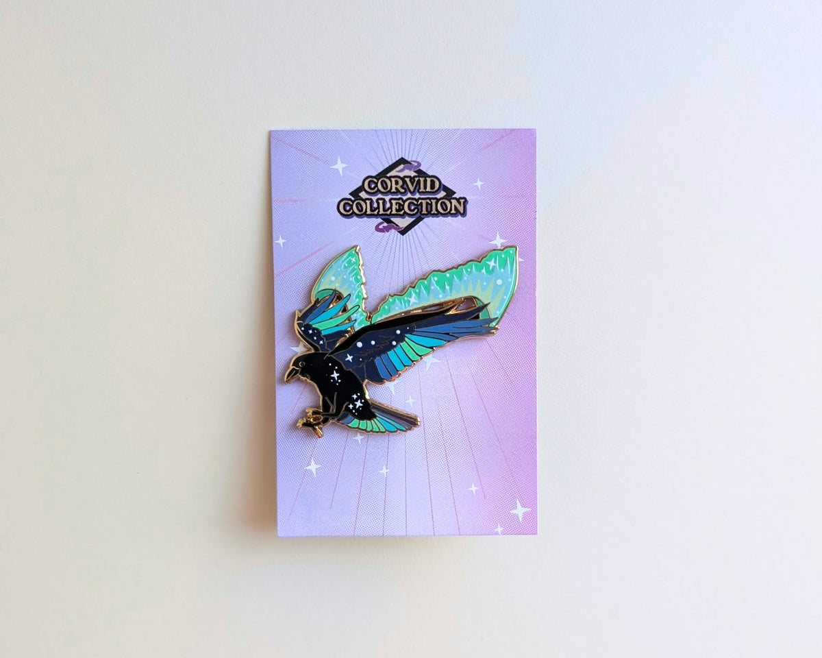 A raven enamel pin with teal, green, and black wings and star details, mounted on a lavender backing card from Glitter Punk's Corvid Collection, displayed against a plain off-white wall.