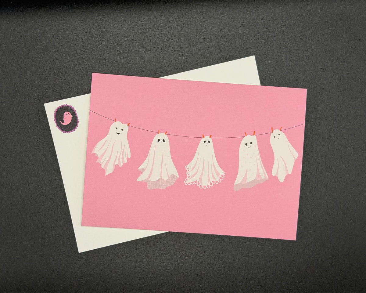 A pink postcard featuring five cute white ghosts hanging on a clothesline.