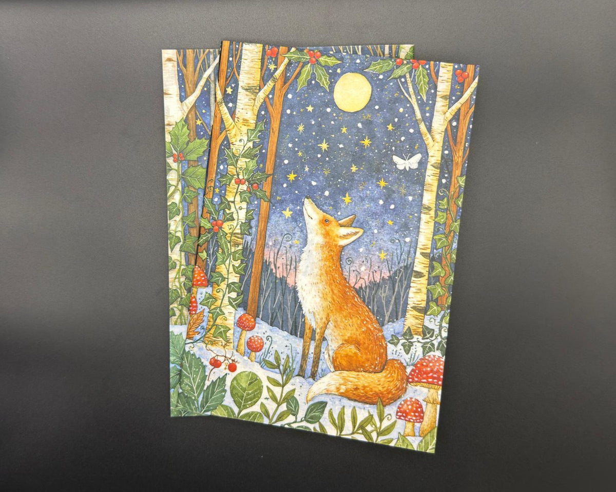 A5 notebook with plain, white pages. The cover features a fox sitting in the snow, looking up at the moon and the holly trees. There are leaves and mushrooms on the floor. 
