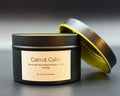 A black metal candle tin labeled 'Carrot Cake' by Thistle Candles, with the lid propped open to show a gold interior, on a dark gray background.