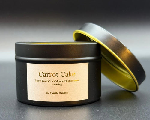 A black metal candle tin labeled 'Carrot Cake' by Thistle Candles, with the lid propped open to show a gold interior, on a dark gray background.