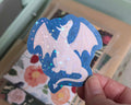 A hand holds a die-cut sticker featuring a white dragon on a dark blue background with holographic star confetti, set against a blurred floral and pastel-colored box.
