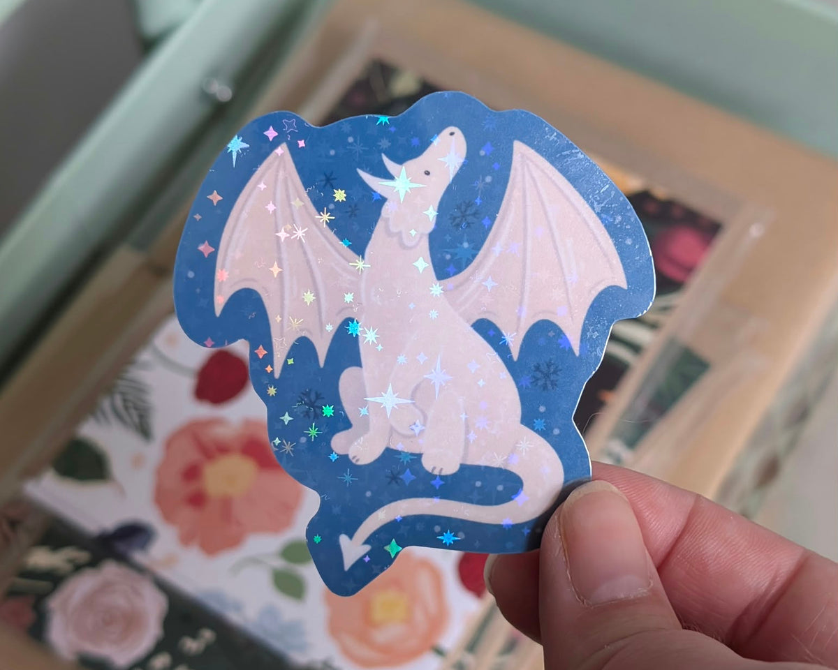 A hand holds a die-cut sticker featuring a white dragon on a dark blue background with holographic star confetti, set against a blurred floral and pastel-colored box.