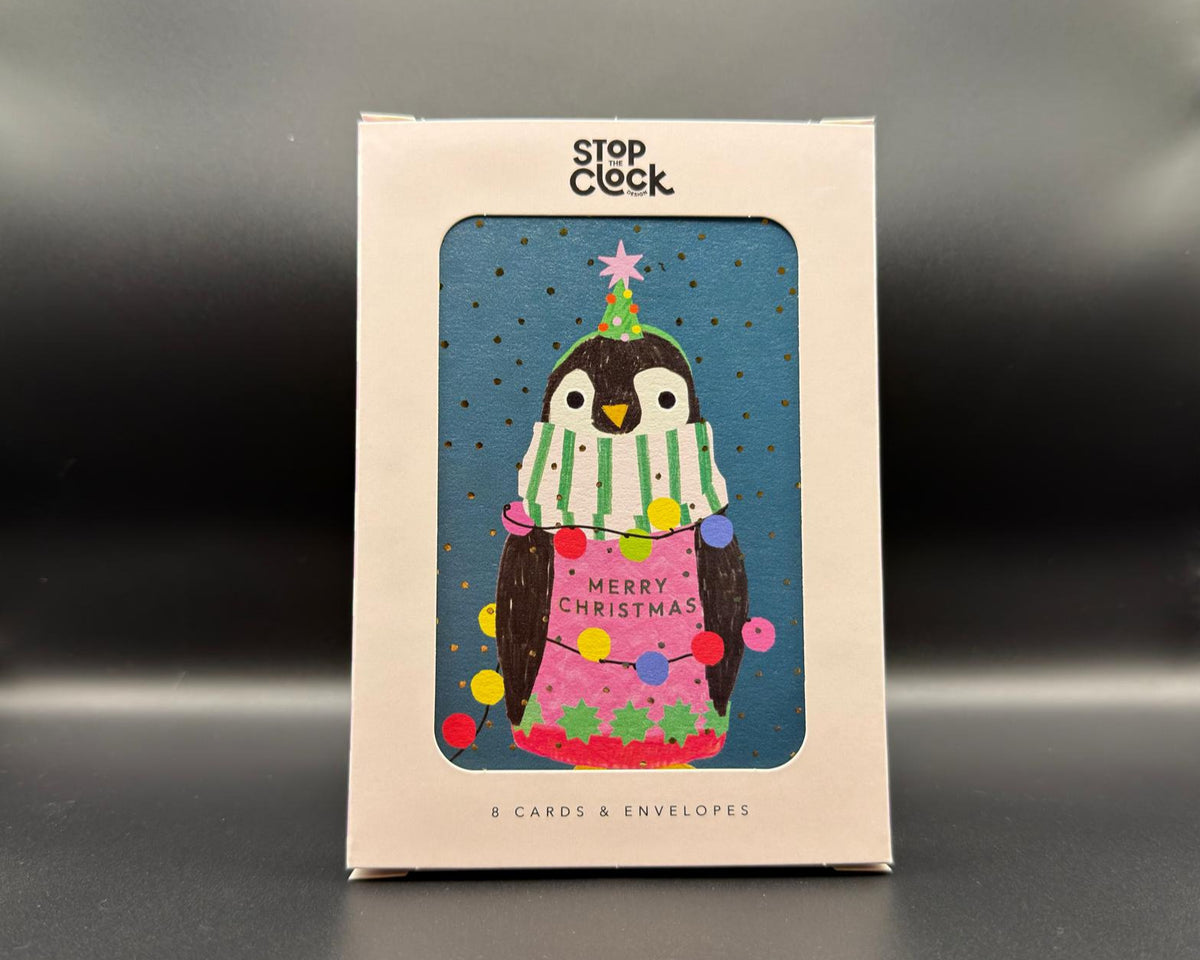 A boxed card set from Stop Cock featuring a penguin illustration on the front. The cream-colored packaging has a clear window showing a blue card with a penguin wearing a green-and-white striped scarf and a pink dress decorated with colourful ornaments. The text says, 'Merry Christmas'. 