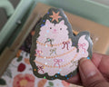A hand holds a scalloped die-cut sticker featuring a fluffy white cat dressed as a Christmas tree with colourful bows and a star topper.