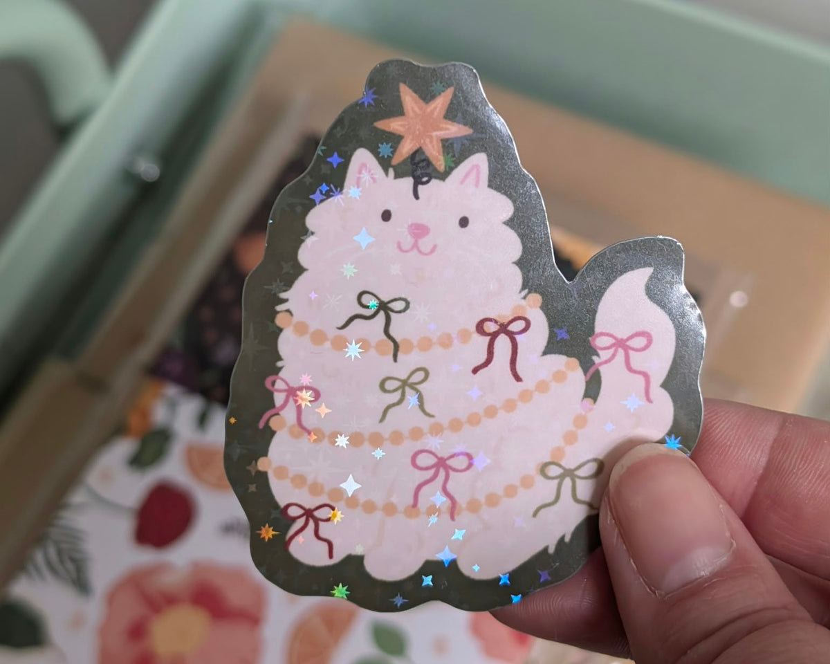 A hand holds a scalloped die-cut sticker featuring a fluffy white cat dressed as a Christmas tree with colourful bows and a star topper.