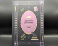 A reading journal with a dark gray cover featuring a central lavender oval, gold decorative border, and occult-themed illustrations including a raven, snake, moon phases, stars, and an eye of providence.