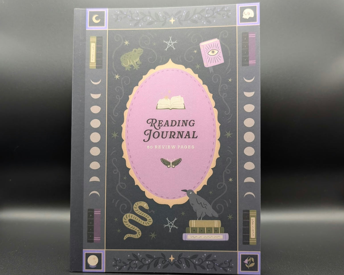 A reading journal with a dark gray cover featuring a central lavender oval, gold decorative border, and occult-themed illustrations including a raven, snake, moon phases, stars, and an eye of providence.