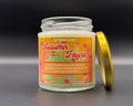 A glass jar candle with a gold lid leaning against it on a dark grey surface. The candle label reads 'The Moodie Store Autumn Fayre Apple Butter Vanilla' with a colourful autumn-themed design in orange, yellow and red.
