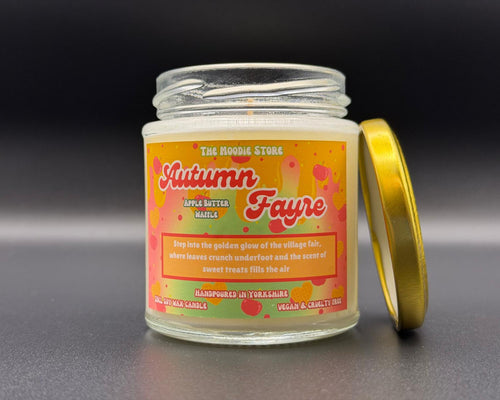 A glass jar candle with a gold lid leaning against it on a dark grey surface. The candle label reads 'The Moodie Store Autumn Fayre Apple Butter Vanilla' with a colourful autumn-themed design in orange, yellow and red.