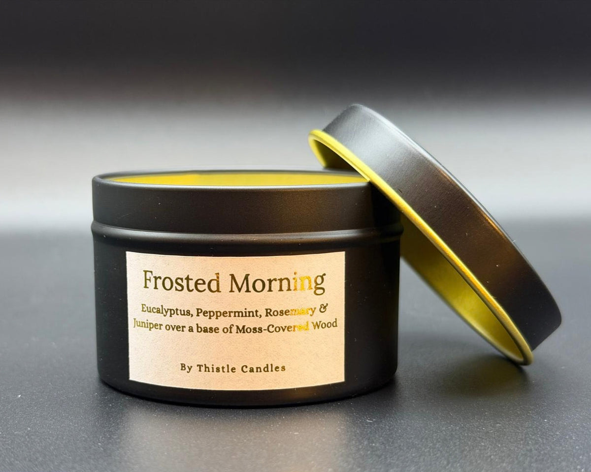 A black metal candle tin with a cream label reading 'Frosted Morning' and 'By Thistle Candles,' shown with the lid propped open against the side. The candle is photographed on a dark gray surface with a soft gray background.