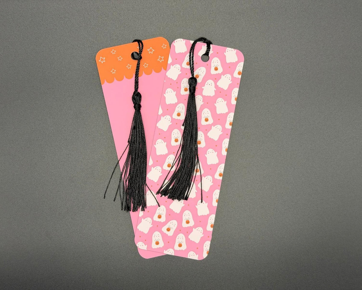 Pink paper bookmark with white ghosts holding orange pumpkins. The bookmark has rounded corners and a black fabric tassel. 