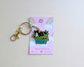 A cute enamel pin of a black and white cat sitting in a green planter, attached to a gold keyring and displayed on its pink backing card by Glitter Punk.
