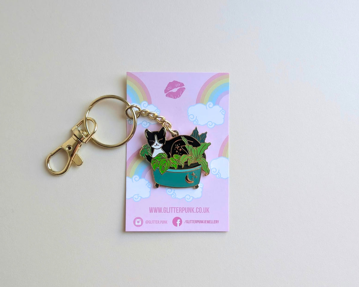 A cute enamel pin of a black and white cat sitting in a green planter, attached to a gold keyring and displayed on its pink backing card by Glitter Punk.