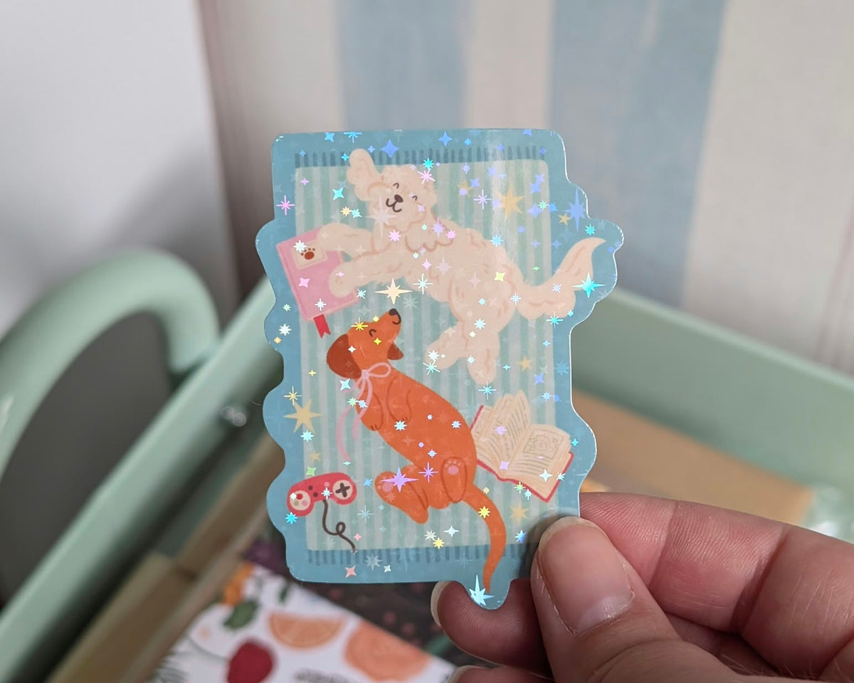 A hand holding a scalloped, die-cut sticker featuring a whimsical illustration of a white dog and an orange dog laying next to pink books and a pink gaming controller on a light blue striped background with holographic star glitter.